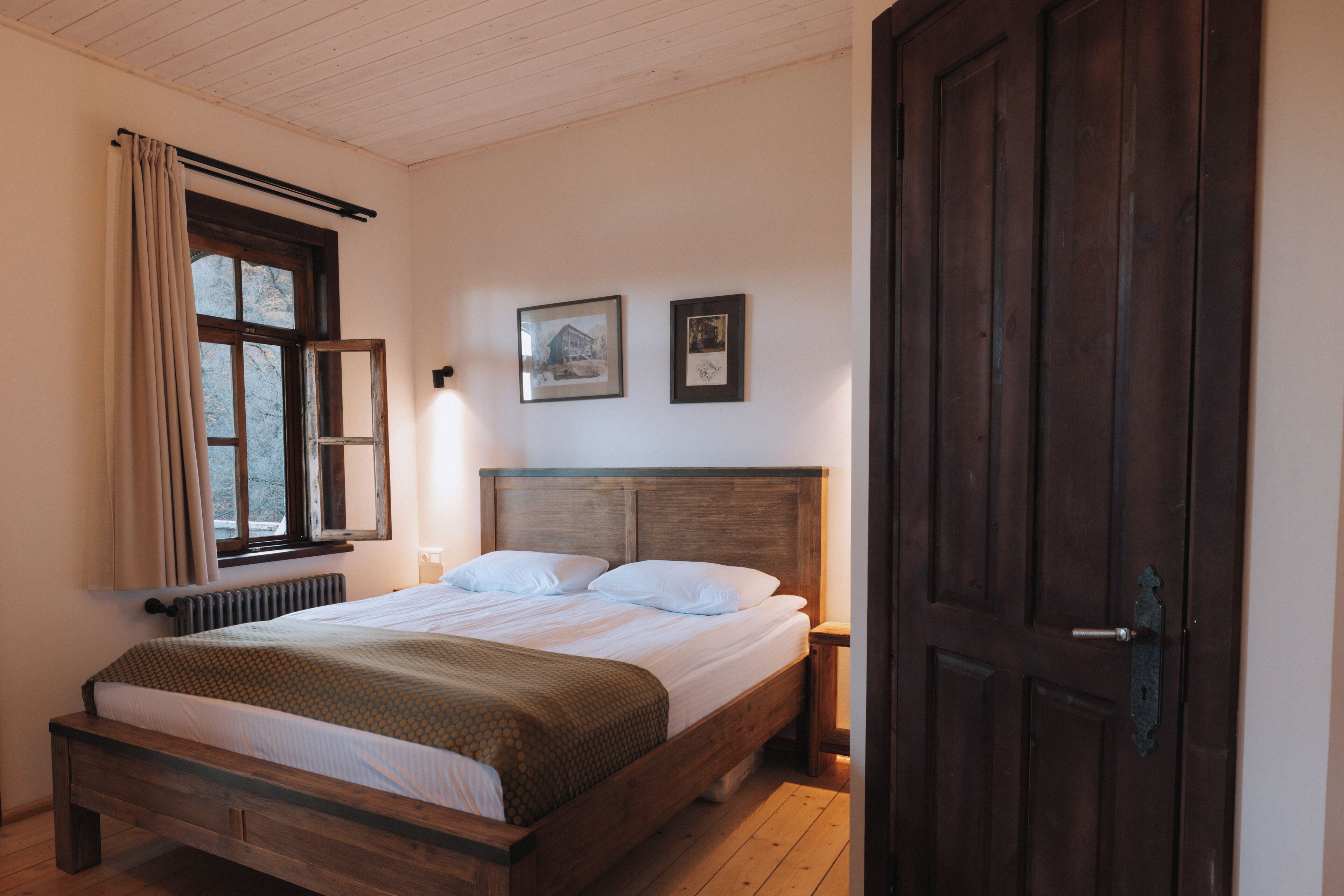 A bedroom with a wooden bed, a window with curtains, a nightstand, and two framed pictures on the wall.