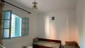 Ein kleiner, schlichter Raum mit zwei Betten, einem Tisch und einem Fenster mit Rollläden.