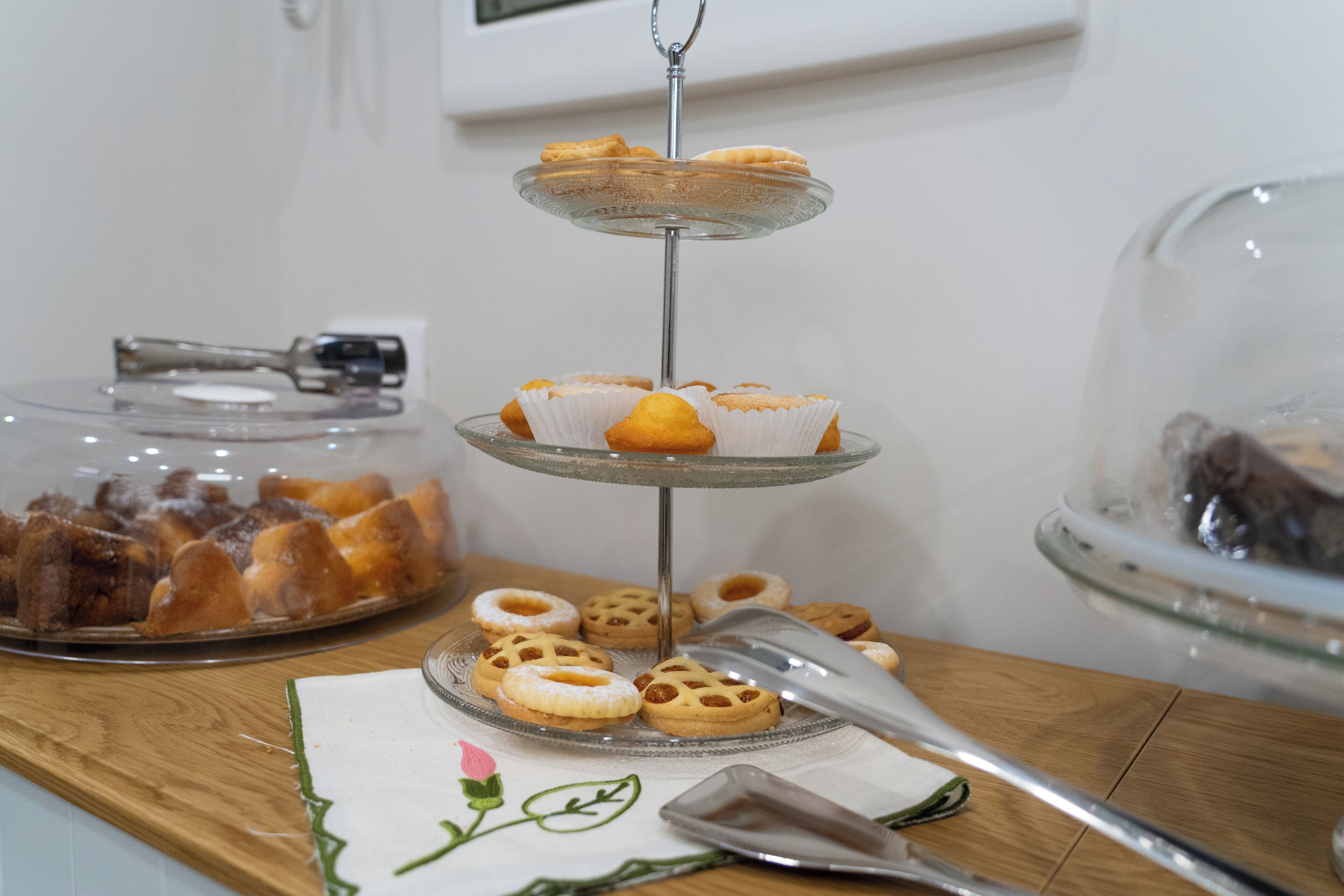 A three-tiered glass dessert stand with pastries, a glass container of bread, and a wooden table with a cloth.