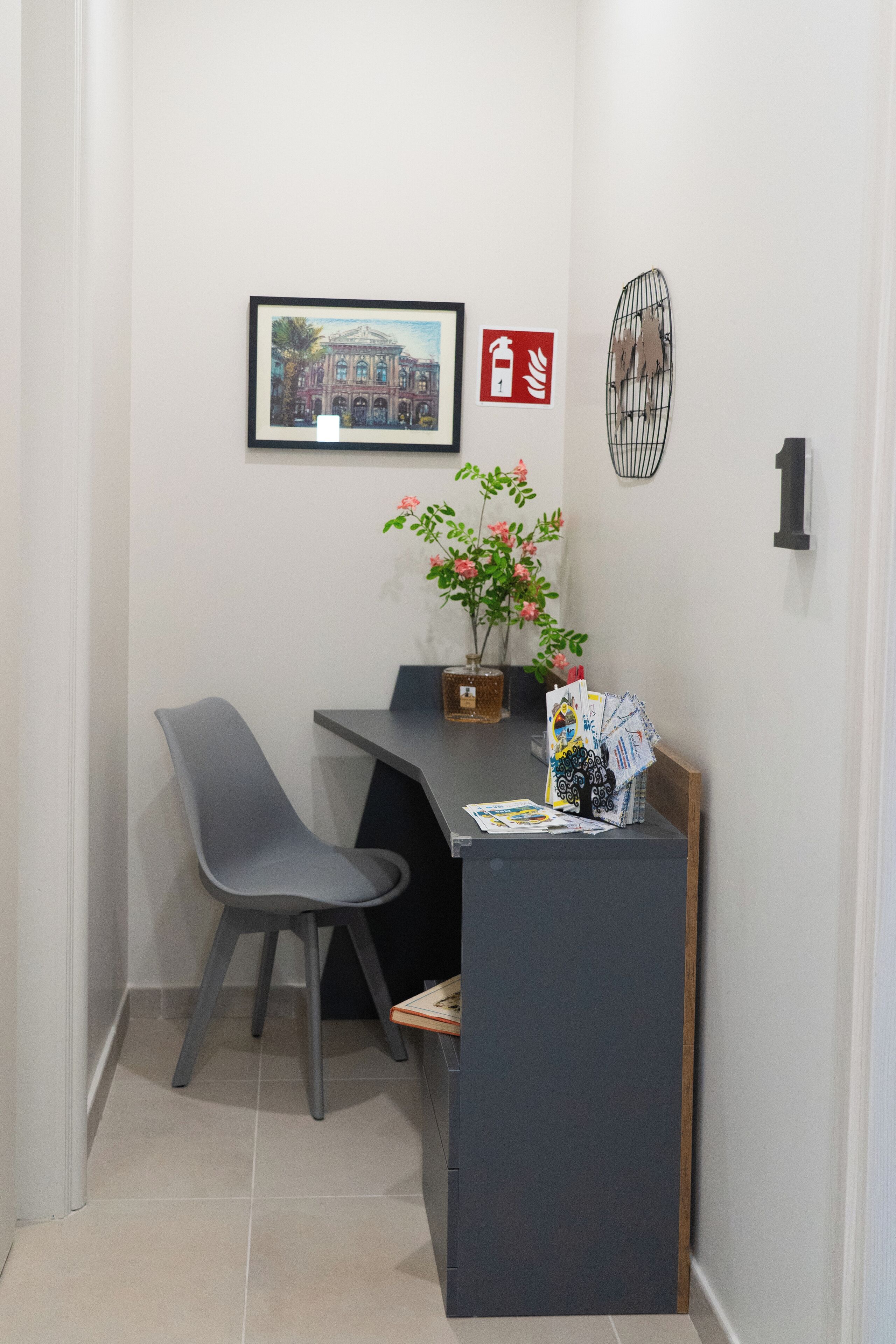 A small, modern room with a desk, two chairs, a potted plant, a framed picture, and a fire exit sign.