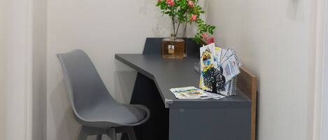 A small, modern room with a desk, two chairs, a potted plant, a framed picture, and a fire exit sign.