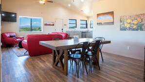 A spacious living area with a red sofa, a dining table with chairs, a kitchen area, and a large window offering a view of the landscape. - Romantic Wine Country Retreat • Private Hot Tub & Valley Views (Cornville)
