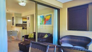 Un espacio de estar con puerta corrediza de vidrio, un sofá, una silla, una zona de comedor con mesa y sillas, y una cocina al fondo.