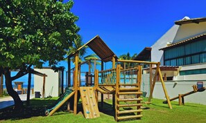 A playground with climbing structures and a slide, surrounded by a building with a white facade and a palm tree. - EKO104 | Cozy beachfront apartment in Muro Alto. (Ipojuca)