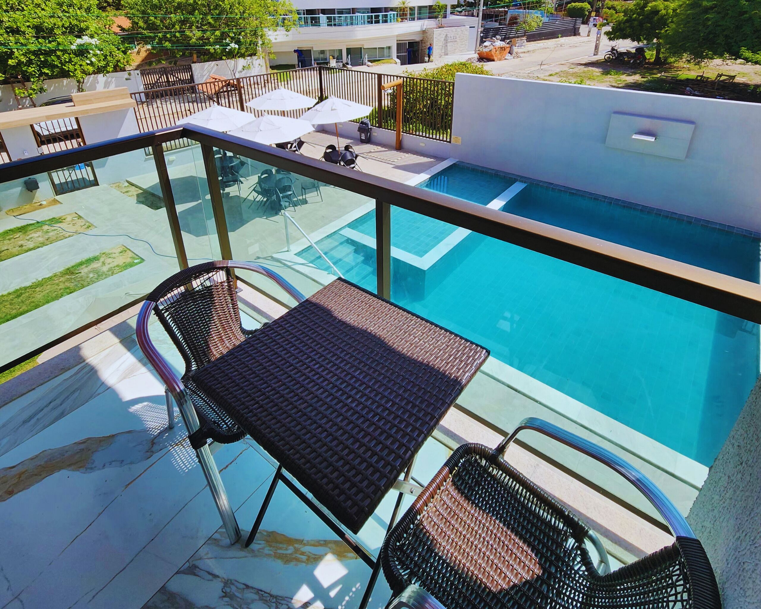 A balcony with a glass railing overlooking a swimming pool, equipped with outdoor furniture and a view of the surrounding area.