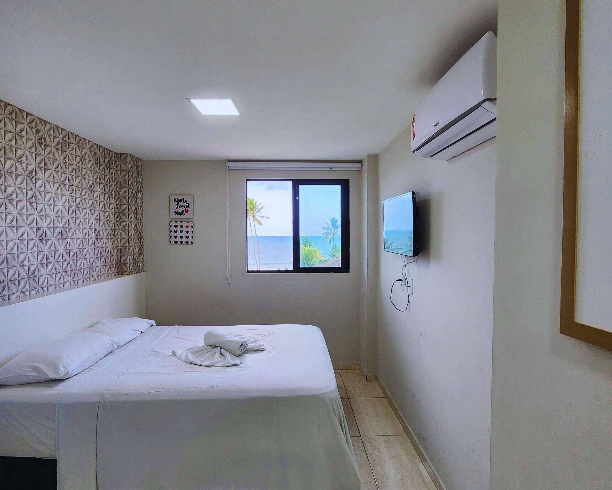 A hotel room with a bed, a TV mounted on the wall, a window with a view of the sea, and a patterned wall.