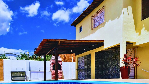 A swimming pool with a brick structure in the background, a covered patio, and a building with patterned windows. - GRA325 | Spacious Retreat with Pool and Barbecue. (Gravatá)