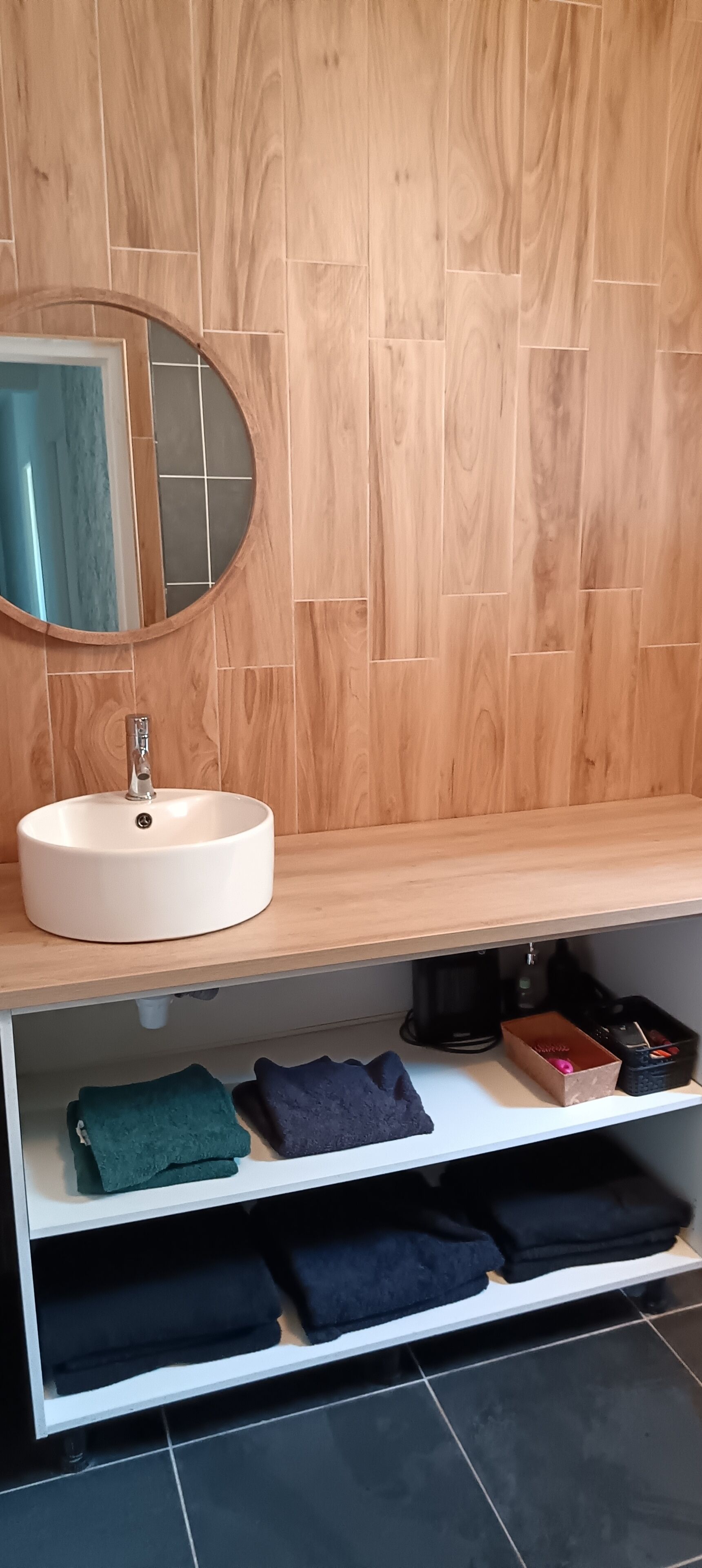 A bathroom with a wooden wall, a round mirror, a white sink, and shelves with folded towels.