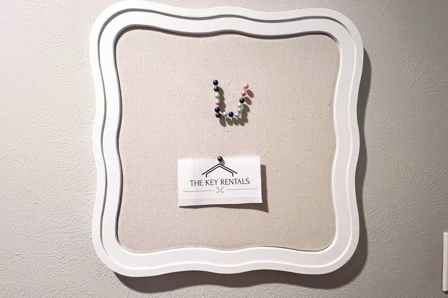 A wall-mounted display with a white decorative frame containing a small sign labeled "THE KEY RENTALS" and a string of colorful beads.
