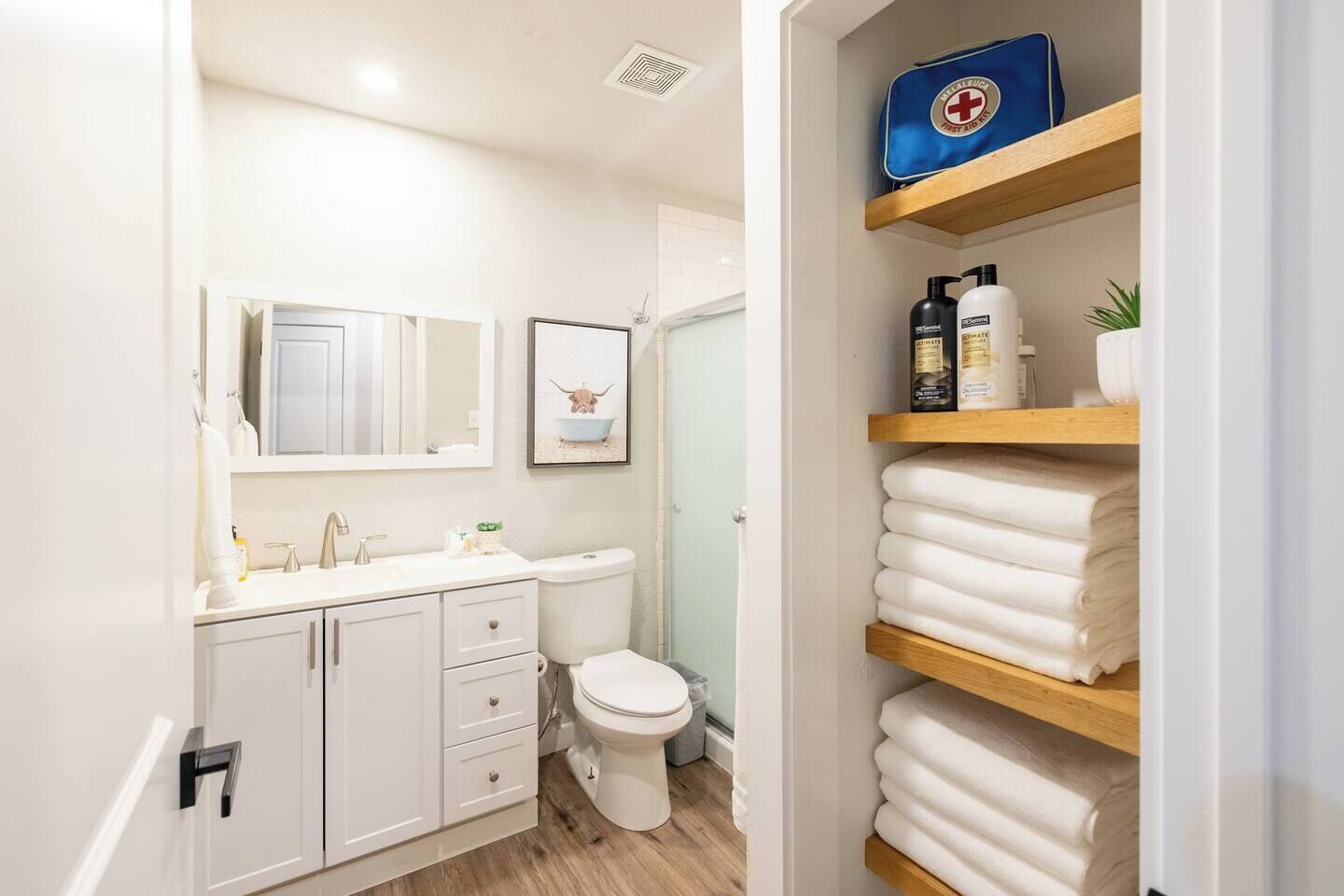 Une salle de bain moderne avec un lavabo blanc, un miroir et des toilettes. On y trouve des étagères avec des articles de toilette et des serviettes.