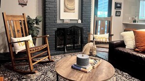 Un salon chaleureux avec une cheminée, un fauteuil à bascule, une table basse avec des livres et un canapé agrémenté d’un coussin moelleux.