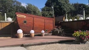 Un bâtiment en bois en forme de bateau, doté d’un petit porche et de deux jardinières contenant des objets sphériques blancs.