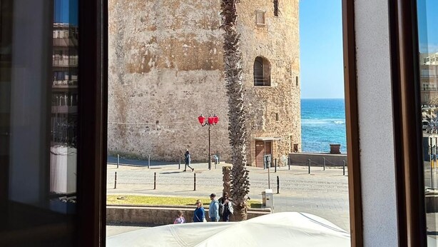 A view of a historic stone building with a palm tree in front, a clear sky, and a body of water in the background. - ALGHERO CENTER LOFT MARIS IN SULIS SQUARE: ICONIC WATERFRONT LOFT-TOP LOCATION (Alghero)