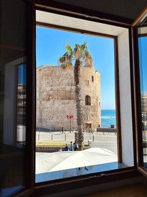 Une vue d’un bâtiment historique en pierre, avec un palmier devant, un ciel dégagé et une étendue d’eau en arrière-plan.