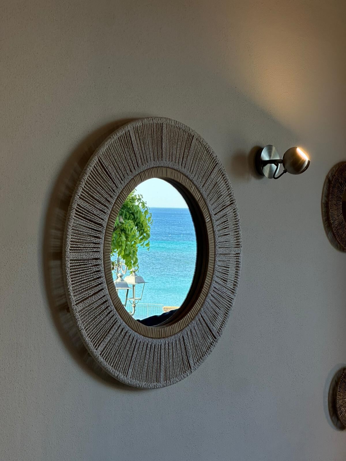 A round mirror with a textured frame on a wall, a wall sconce, and a partial view of a blue ocean through the mirror.