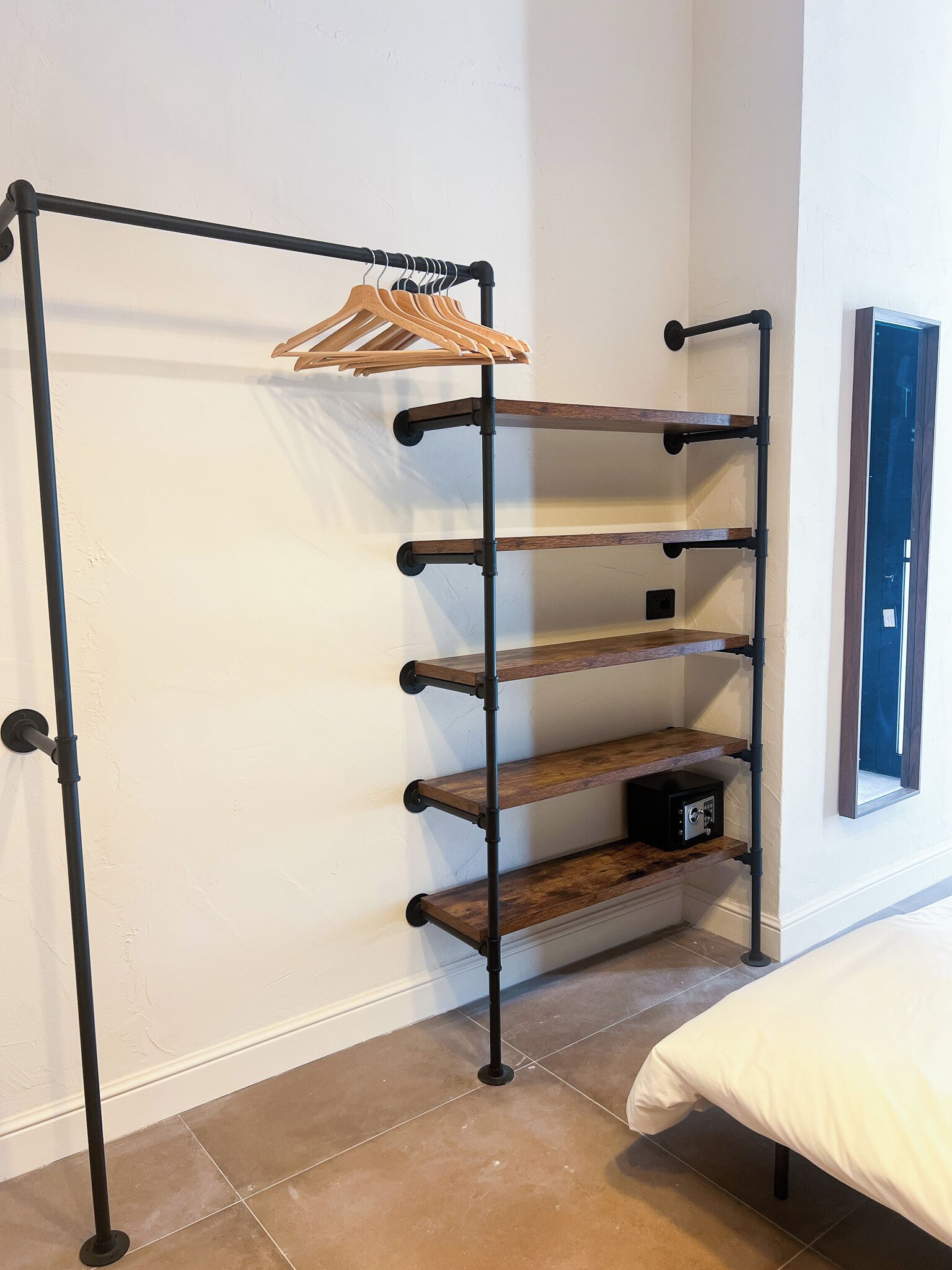 A corner room with a black metal rack holding wooden hangers and shelves, a bed, and a door.