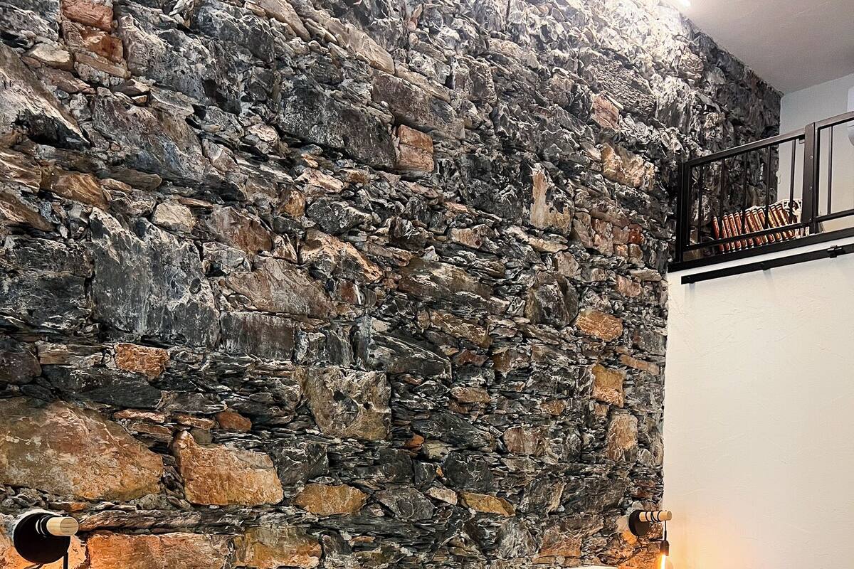 Un dormitori amb pared de pedra, estructura de llit de fusta i una tauleta de nit amb una làmpada.
