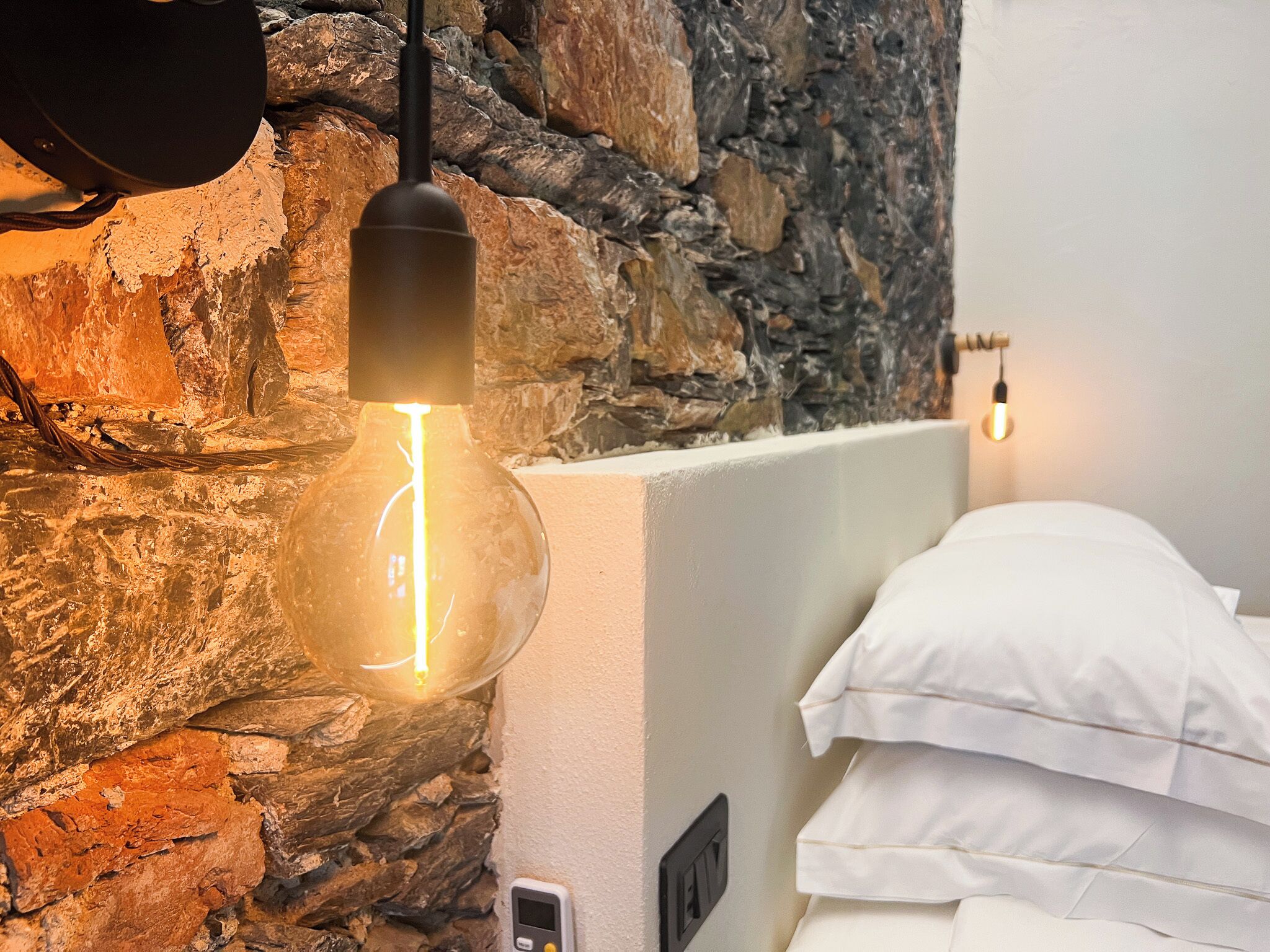 Ein Zimmer mit Steinwand, einem Bett mit weißer Bettwäsche und einer hängenden Deckenleuchte.