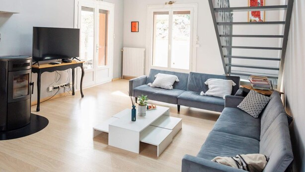 Una sala de estar moderna con sofá, una mesa de centro, televisor y chimenea. Se ve una escalera con barandilla y tragaluz.