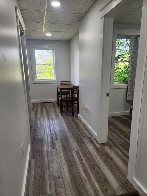 A hallway with wooden flooring, a dining table with chairs, and a window with a view of greenery. - Ninth Door Mention Apt 13 Main St. Milbridge (Milbridge)