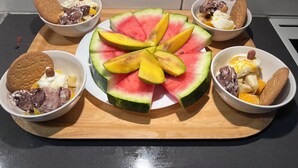 Una bandeja de madera con una rebanada grande de sandía en el centro, rodeada de recipientes con helado, galletas y nueces.
