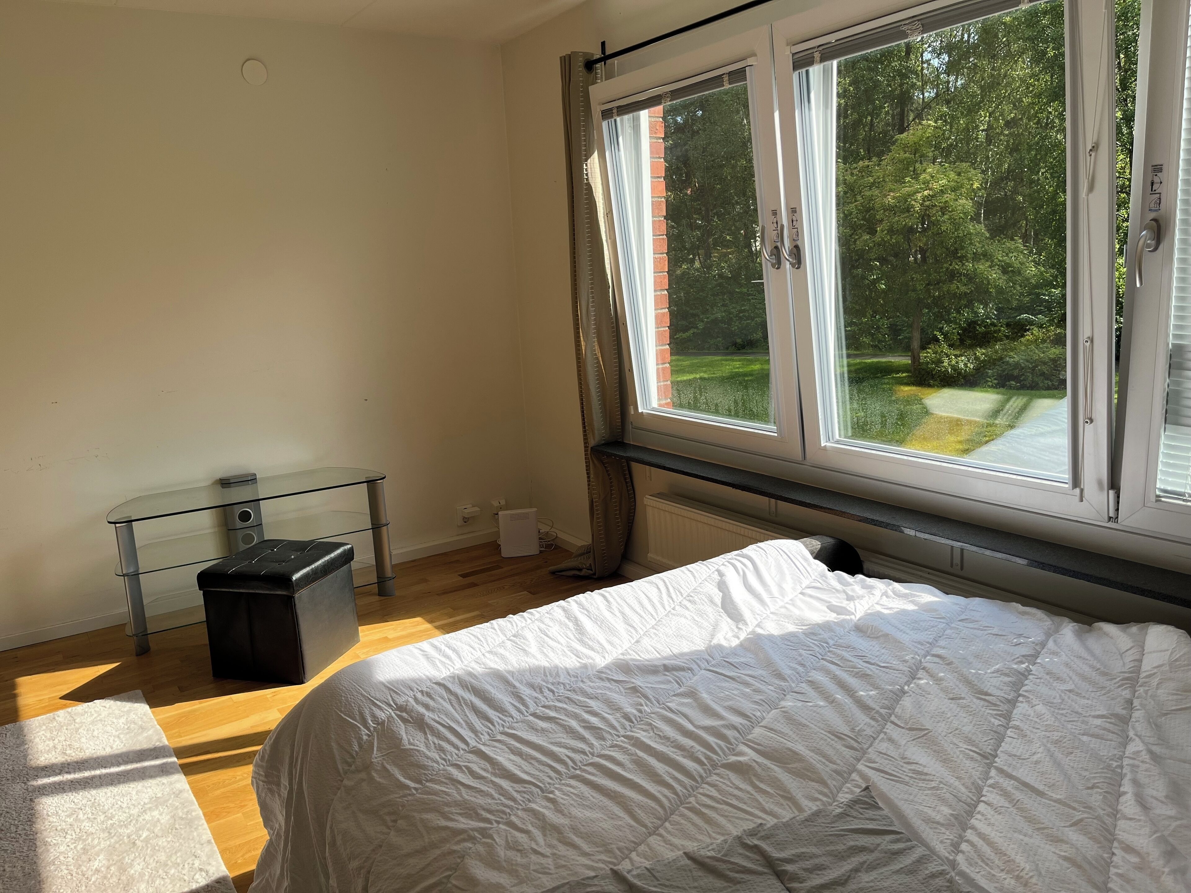 Une chambre avec une grande fenêtre, un lit, une table en verre et un radiateur.