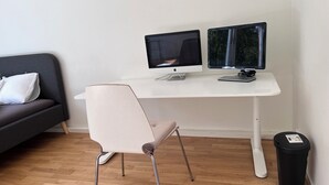 Una habitación moderna con un escritorio blanco, dos monitores, una silla gris, un sofá gris y un cesto de basura negro.