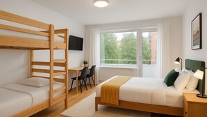 Ein Schlafzimmer mit Etagenbett, Schreibtisch mit zwei Stühlen, Bett mit grünem Kopfteil, Nachttischlampe und Fenster mit Blick auf Bäume und Gebäude.