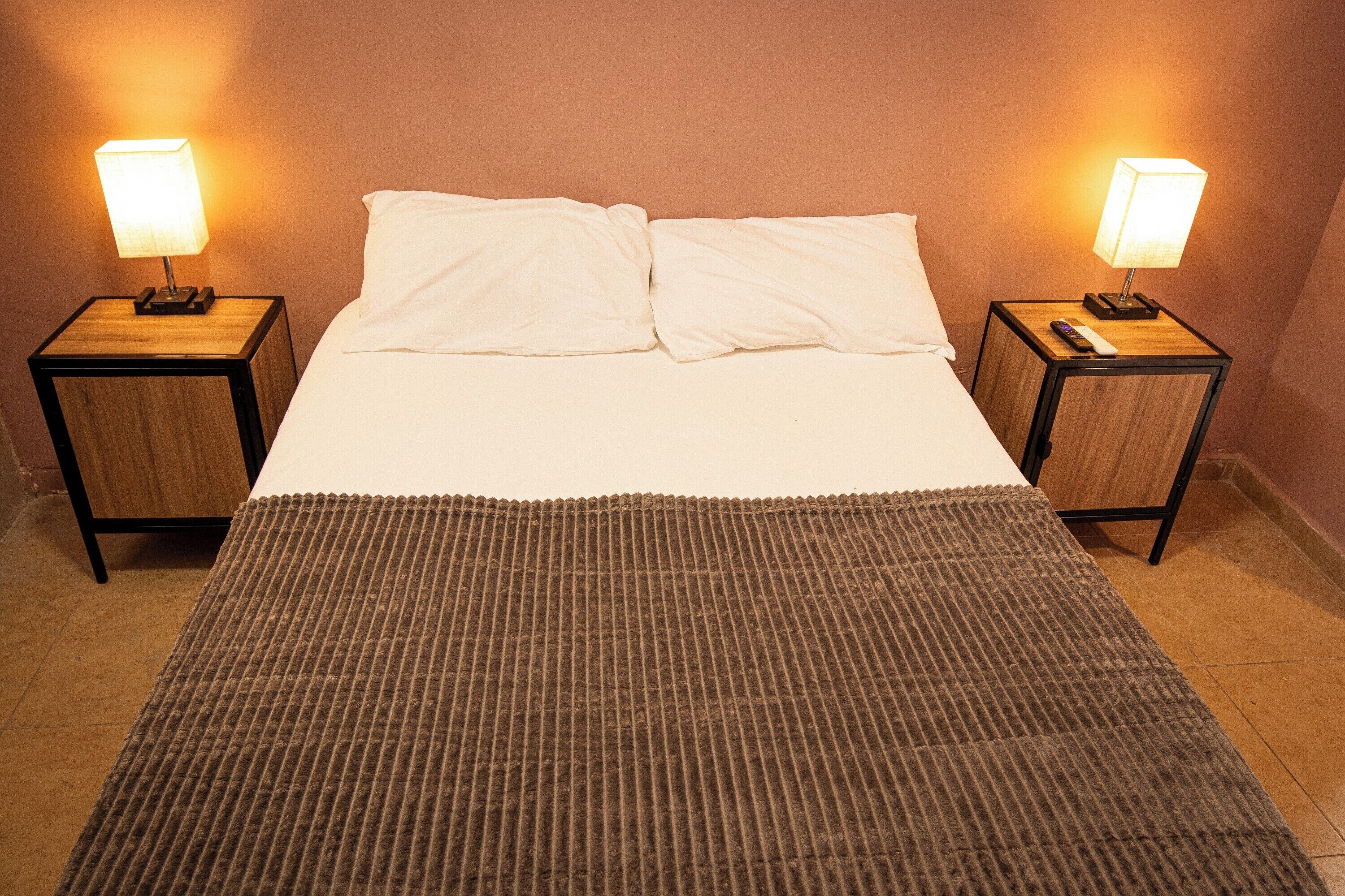 Cama doble con dos mesitas de noche, cada una con una lámpara, en una habitación con un tapete marrón texturizado.