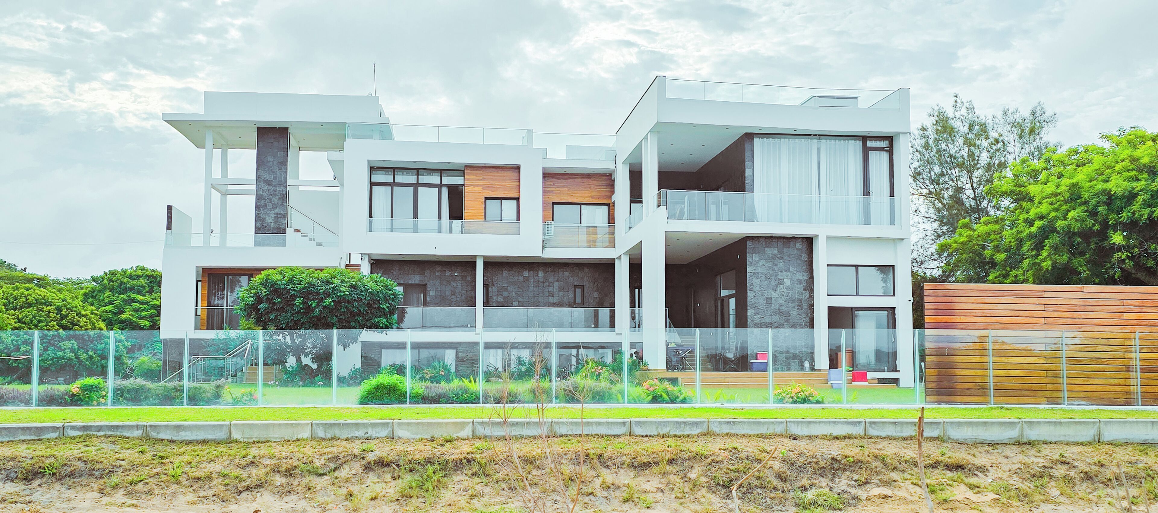 Una casa moderna de varios pisos con balcón de cristal, terraza de madera y jardín cercado.