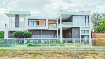 Una casa moderna de varios pisos con balcón de cristal, terraza de madera y jardín cercado.