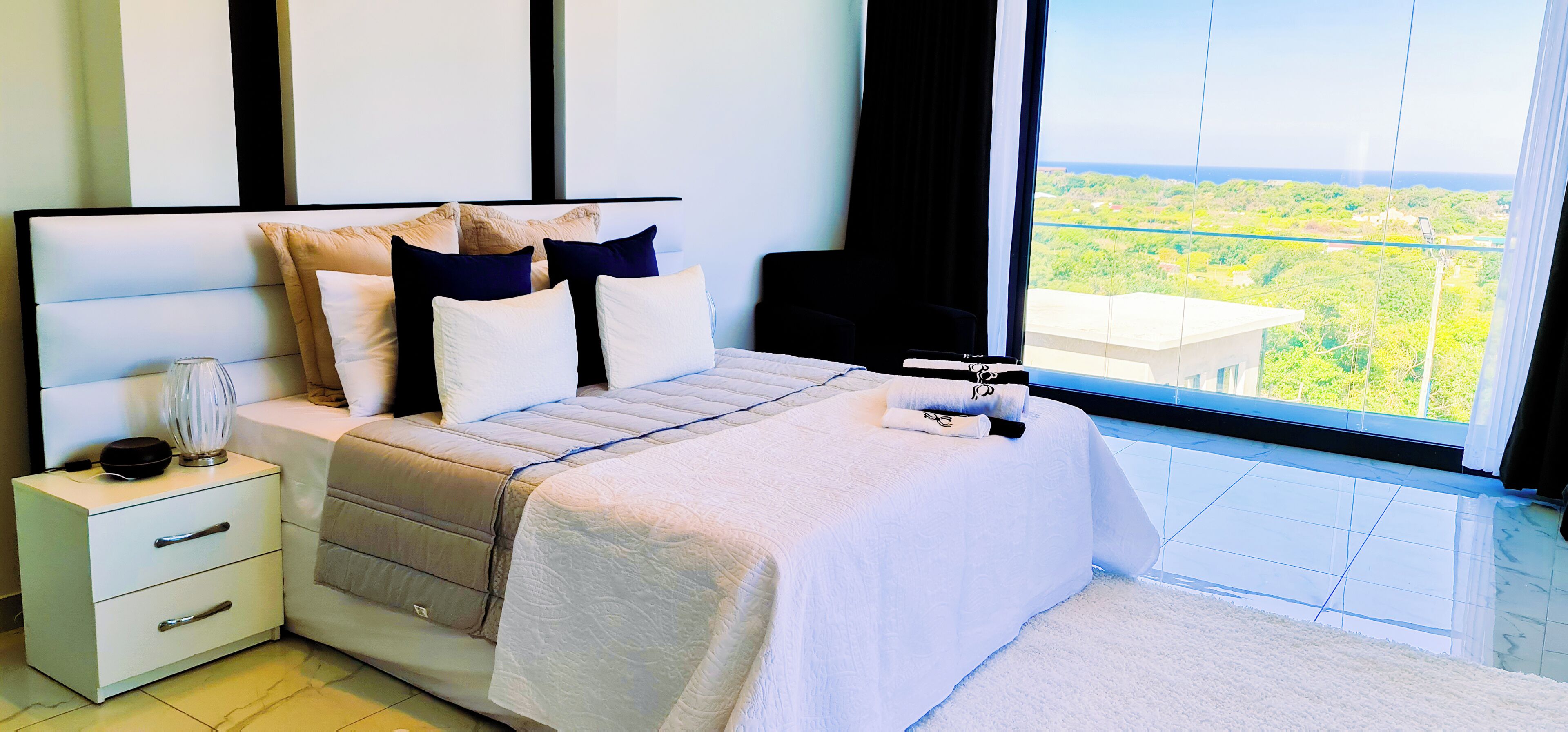 A bedroom with a bed, a nightstand, a lamp, and a view of greenery through the window.