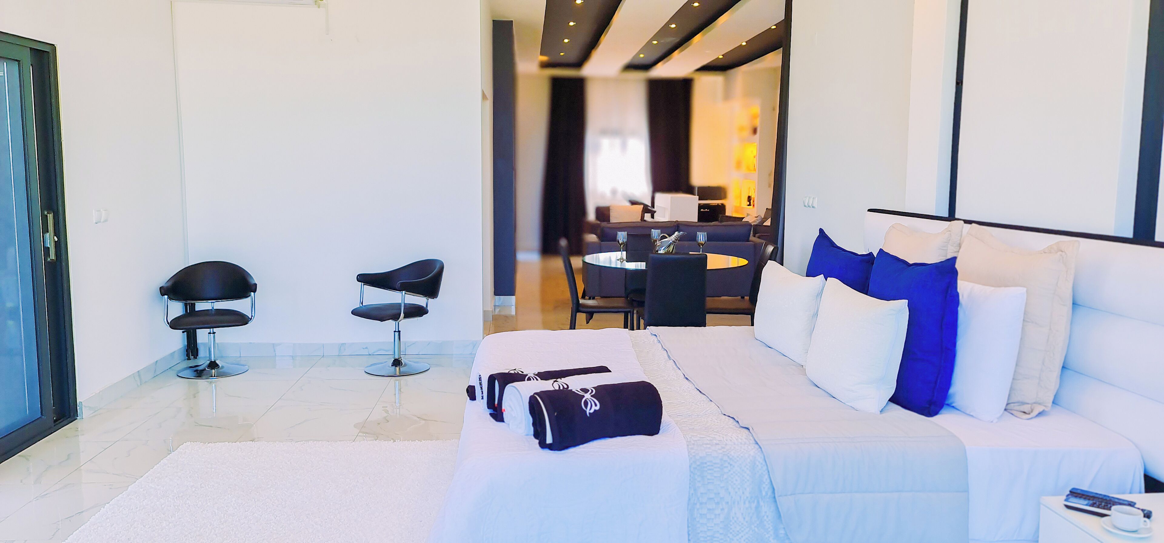 A modern hotel room with a large bed, two black chairs, a dining table with chairs, and a small table with a cup and remote control.