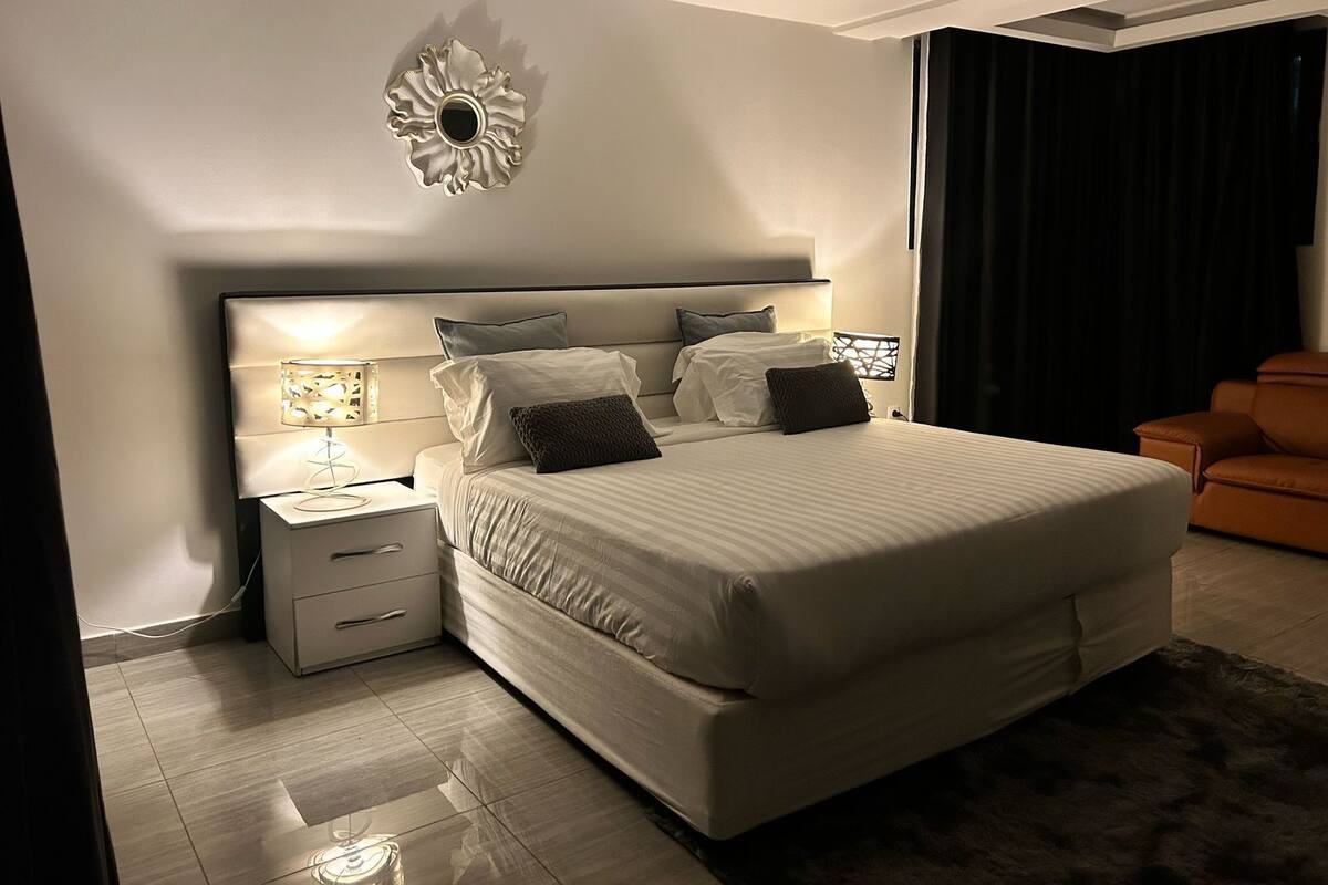 Un dormitorio moderno con una cama grande, mesitas de noche, un adorno en la pared y una cortina oscura.