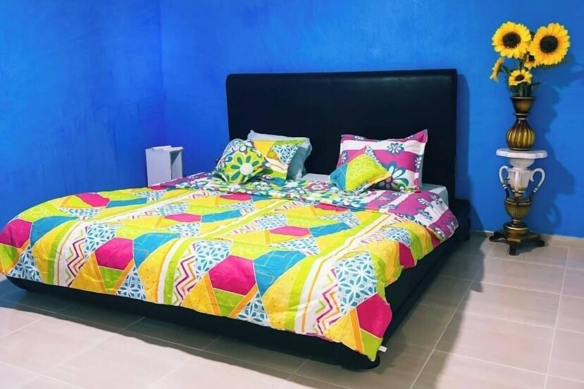 Un dormitorio con cama, paredes azules, aire acondicionado, un florero con girasoles y una mesita.