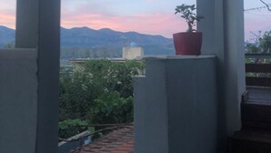 Vista desde un balcón con una planta en maceta, que da a un paisaje urbano con montañas al fondo y un cielo despejado.