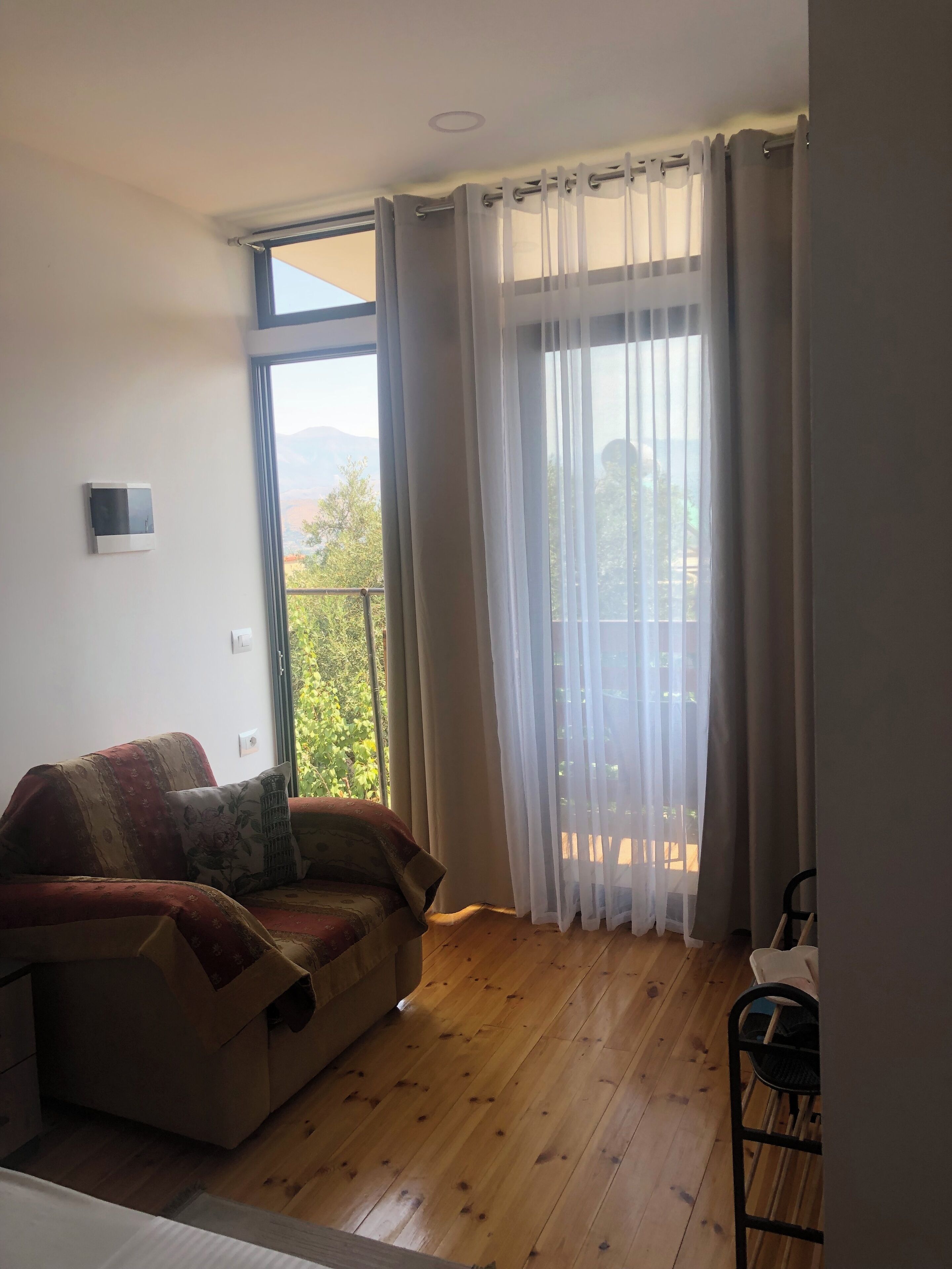 A room with a wooden floor, a sofa with a floral cushion, a window with sheer curtains, and a view of greenery and mountains.