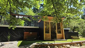Une cabane moderne en bois, dotée d’une structure vitrée, entourée d’un jardin et d’arbres.