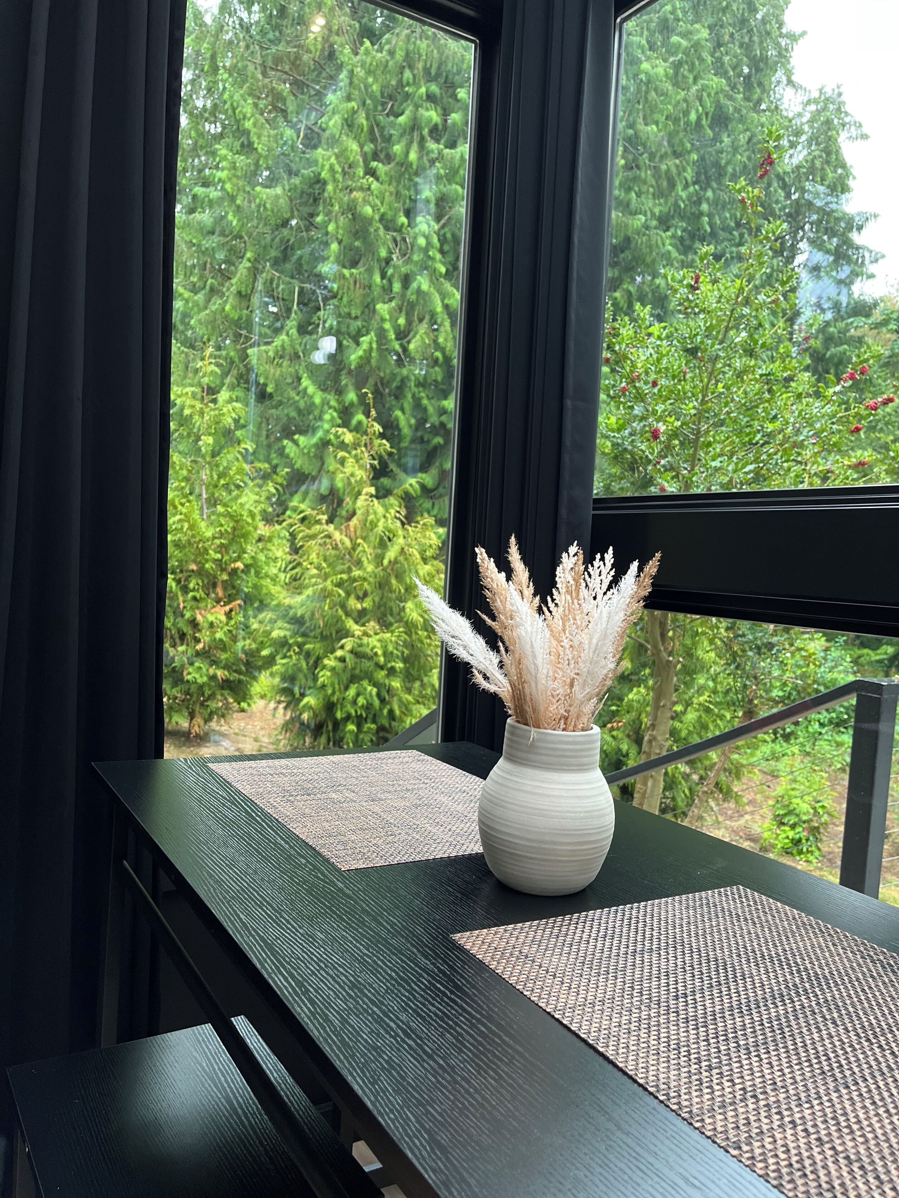 A view through a window with a white vase of dried plants on a black table.