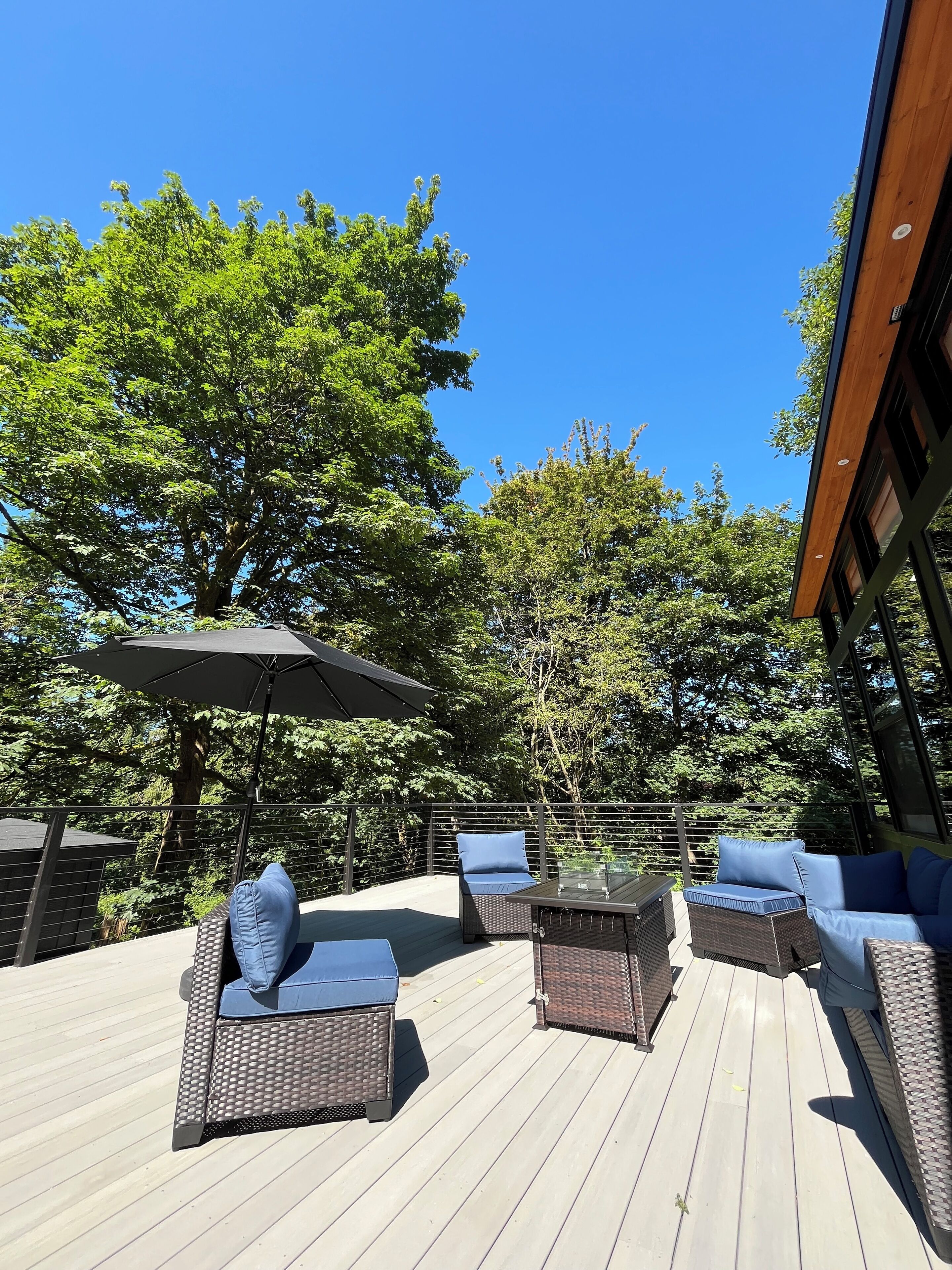 Una terraza de madera con mobiliario de mimbre y una mesa de vidrio, rodeada de árboles y un cielo azul despejado.