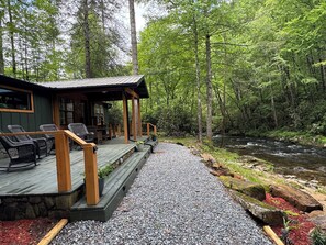 A cabin with a wooden deck, outdoor seating, and a view of a forest and stream.