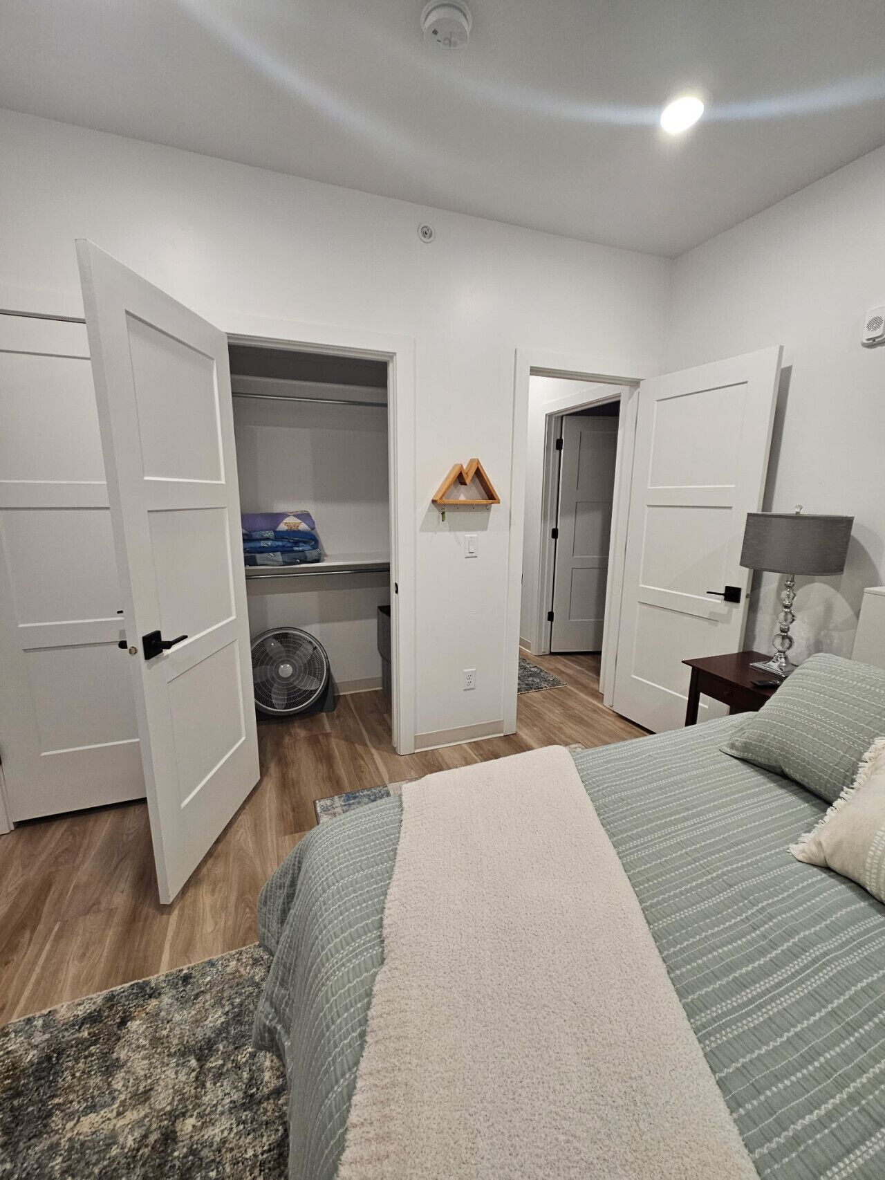 Une chambre à coucher avec un lit, une table de chevet avec une lampe, un ventilateur et un placard ouvert avec des étagères.