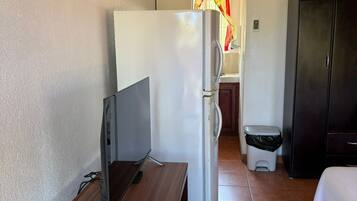 Habitación de hotel pequeña con escritorio de madera, refrigerador blanco, armario negro y cama con ropa de cama blanca.