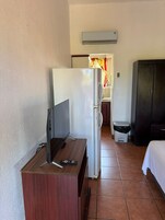 Habitación de hotel pequeña con escritorio de madera, refrigerador blanco, armario negro y cama con ropa de cama blanca.