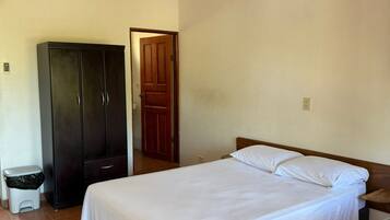Un dormitorio con una cama, un armario, un ventilador de techo, una puerta y un cubo de basura.