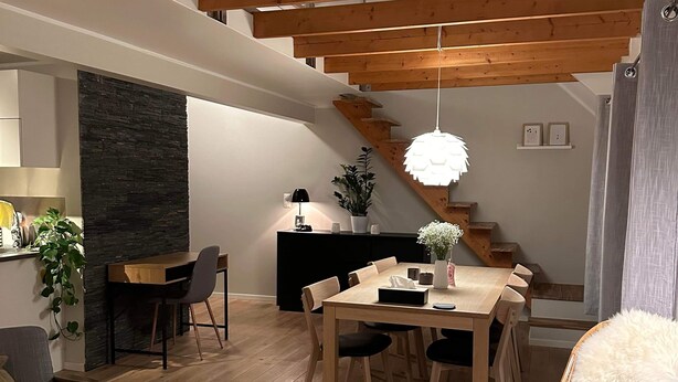 Een ruime woonkamer met een houten eettafel, een bank en een rieten stoel. Een trap met houten balken leidt naar een hoger gelegen verdieping.