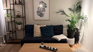 Een moderne woonkamer met een houten salontafel, een zwarte bank met blauwe kussens, een zwart metalen schap met planten, en een kunstwerk in een lijst aan de muur.