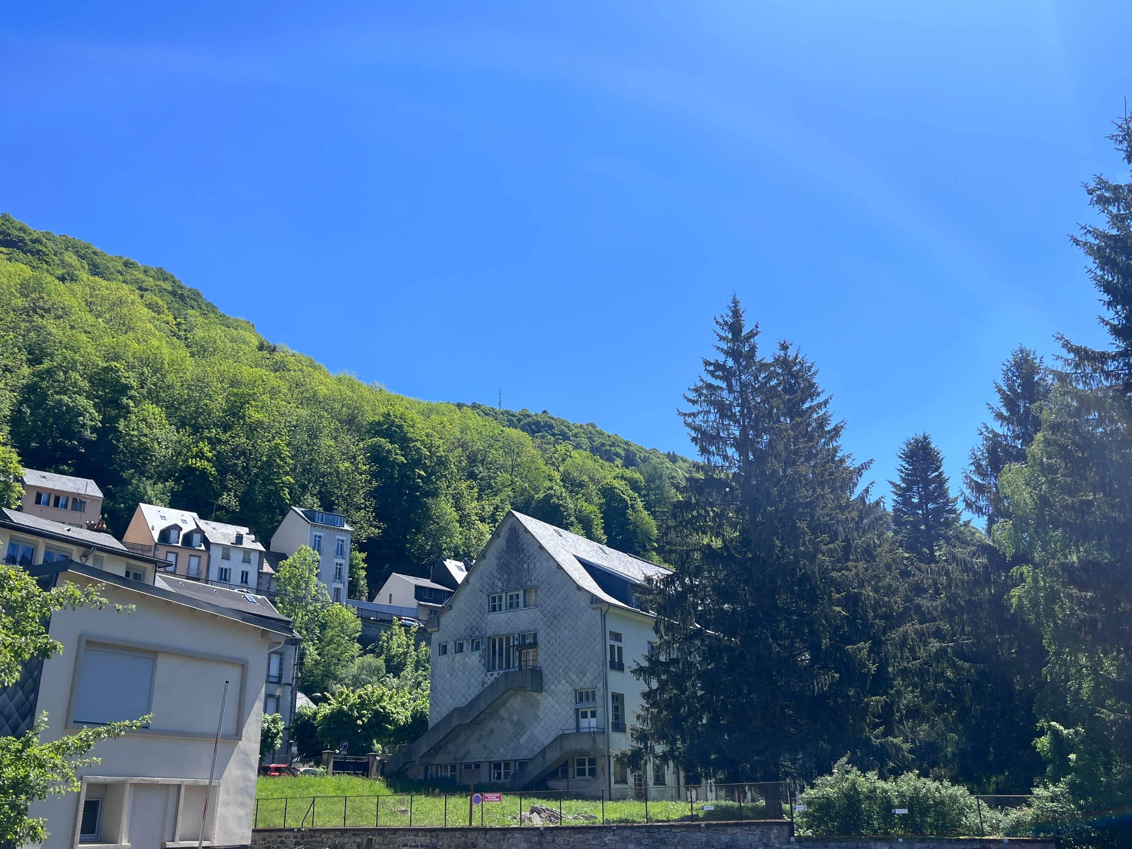 Um edifício com fachada de pedra, várias janelas e telhado inclinado, cercado por vegetação exuberante e árvores.