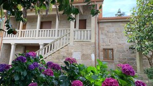 A stone building with a balcony, surrounded by lush greenery and purple flowers.