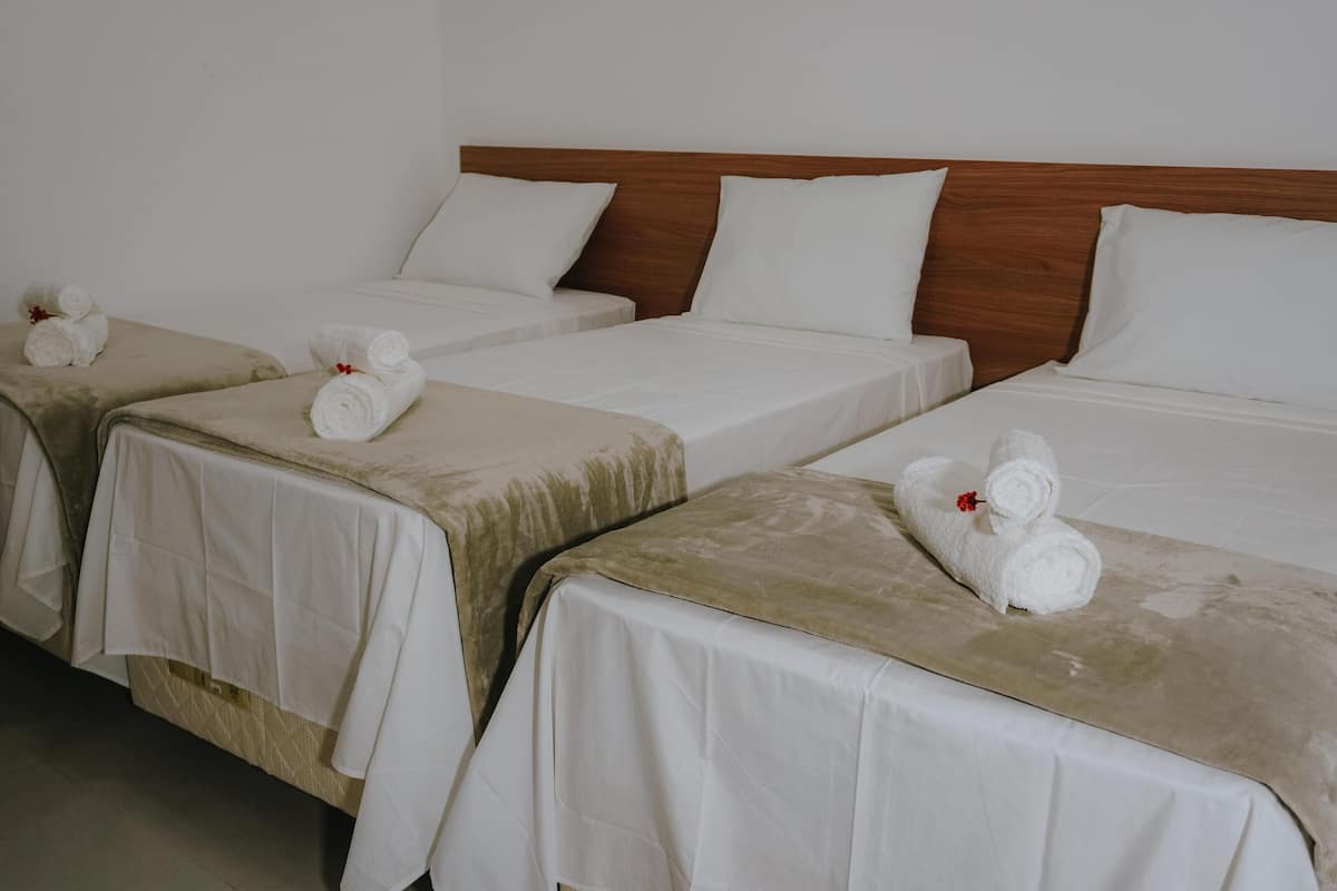 A hotel room with three single beds, each with a white bedspread and a rolled towel with a red flower.